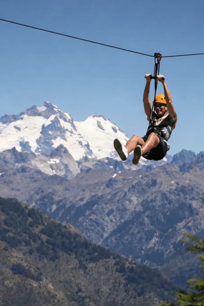 Un hombre con casco naranja se desliza en una tirolesa a gran altura, con un impresionante paisaje de montañas escarpadas y picos nevados bajo un cielo azul despejado.